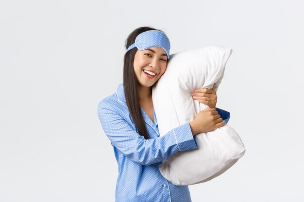 Smiling cute asian girl in sleeping mask and pajamas hugging pillow, like sleeping, feeling pleased as going bed, feeling comfortable and satisfied over white background