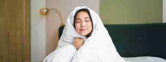 Free photo smiling cute asian woman hugs her warm and cozy duvet feels warm makes cocoon from blanket sits in