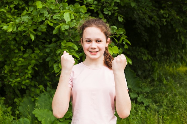 Smiling cute girl clenching her fist making yes gesture while standing in park