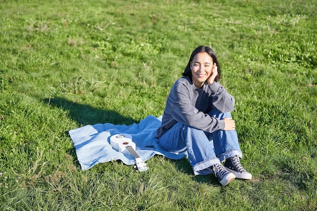 야외에서 화창하고 따뜻한 날씨를 즐기며 우쿨렐레를 연주하는 공원에서 담요에 앉아 웃는 귀여운 소녀