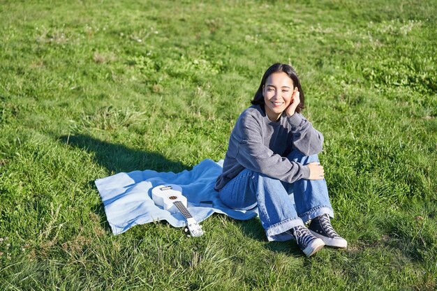 야외에서 화창하고 따뜻한 날씨를 즐기며 우쿨렐레를 연주하는 공원에서 담요에 앉아 웃는 귀여운 소녀