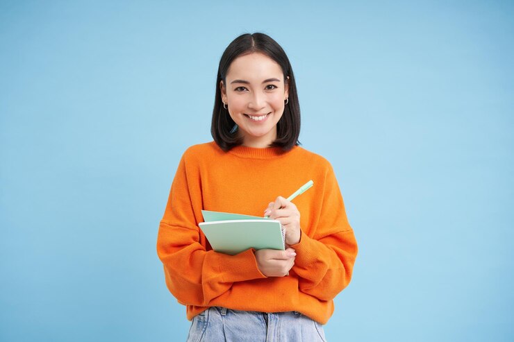 Free Photo smiling cute girl student making notes writing down homework studying standing over blue studio back