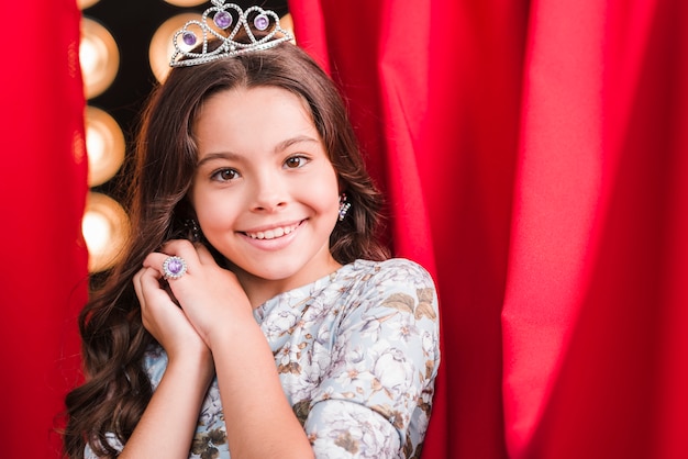 Smiling cute girl wearing crown standing in front of red curtain