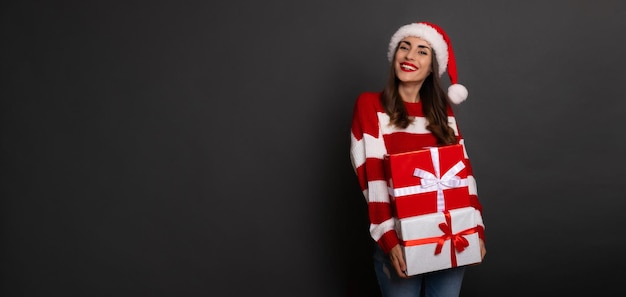 Smiling cute happy young woman in santa hat with the bright colorful present box in hands is having fun while posing isolated in studio