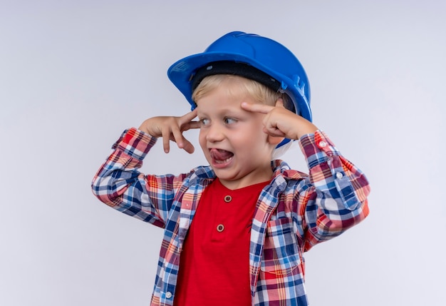 A smiling cute little boy with blonde hair wearing checked shirt in blue helmet pointing at his head with index finger on a white wall