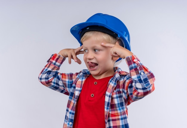 흰 벽에 검지 손가락으로 그의 머리를 가리키는 파란색 헬멧에 체크 셔츠를 입고 금발 머리를 가진 웃는 귀여운 어린 소년