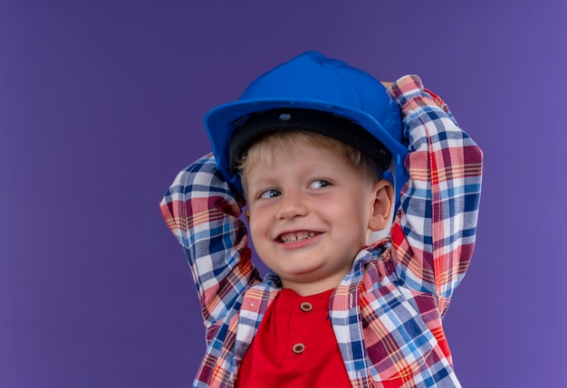 A smiling cute little boy with blonde hair wearing checked shirt holding hand on blue helmet on a purple wall