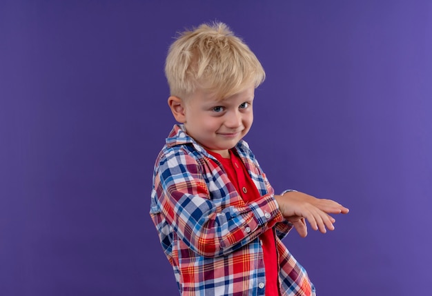 Улыбающийся милый маленький мальчик со светлыми волосами в клетчатой рубашке, подняв руки вверх, глядя на фиолетовую стену