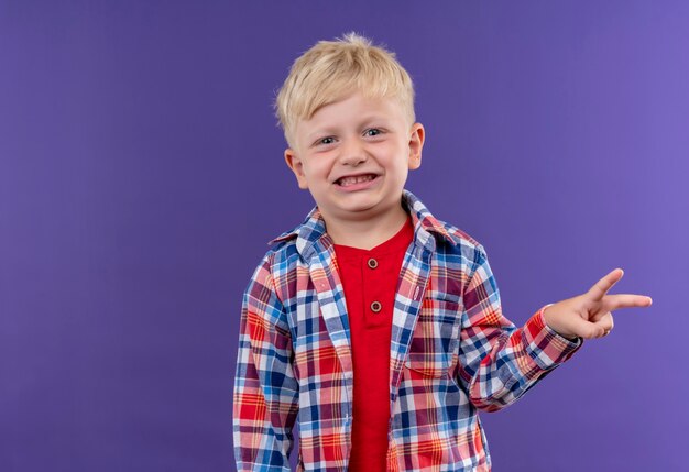 Улыбающийся милый маленький мальчик со светлыми волосами в клетчатой рубашке показывает жест двумя пальцами на фиолетовой стене