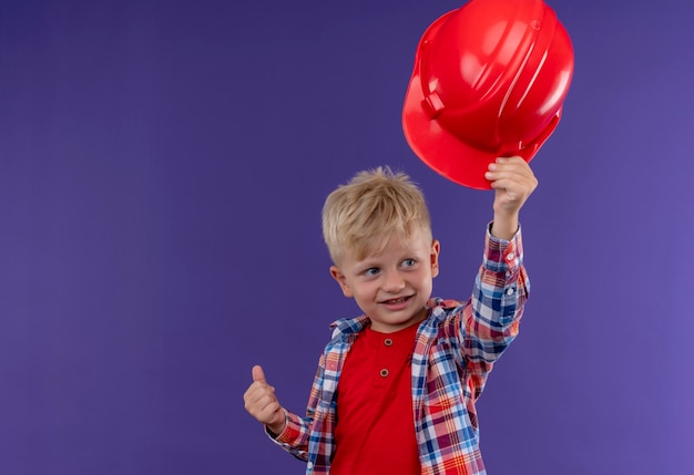 Un ragazzino sveglio sorridente con capelli biondi che indossa l'elmetto rosso di decollo controllato mentre osserva il lato su una parete viola