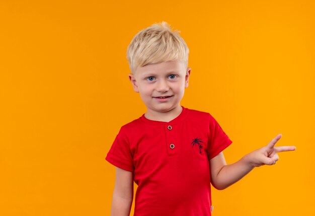 黄色の壁に2本の指のジェスチャーを示す赤いTシャツを着ているブロンドの髪の笑顔のかわいい男の子