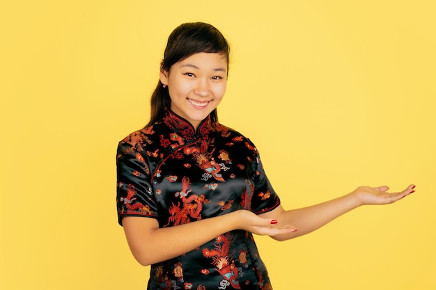 Smiling cute, showing at side. Happy Chinese New Year. Asian young girl's portrait on yellow background. Female model in traditional clothes looks happy. Celebration, human emotions. Copyspace.