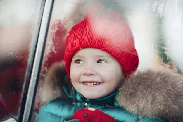 Smiling cute winter girl in red hat sitting in car having fun medium shot. Happy beautiful female baby in warm clothing having positive emotion outdoor surrounded by snowflakes enjoying childhood