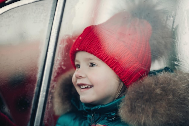 Улыбается милая зимняя девушка в красной шляпе, сидя в машине, весело средний выстрел. Счастливый красивый ребенок женского пола в теплой одежде, имеющий положительные эмоции на открытом воздухе в окружении снежинок, наслаждающийся детством