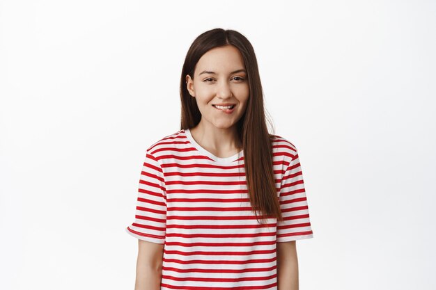 Smiling cute woman biting lip and squinting from tempation, want to try delicious thing, tempted with something, standing in striped t-shirt against white wall.