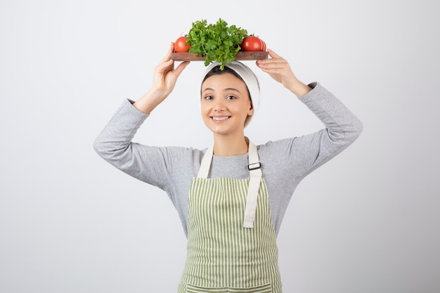 smiling cute woman model holding a wooden board with fresh vegetables on head.