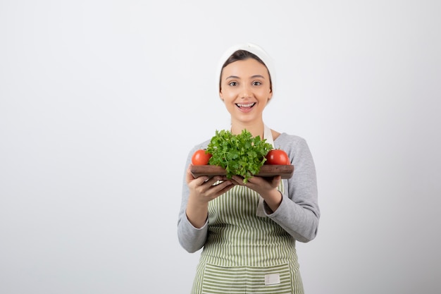 smiling cute woman model holding a wooden board with fresh vegetables .