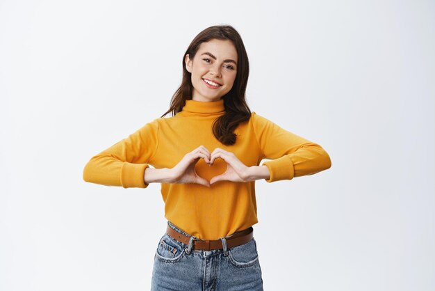 Smiling cute woman showing heart sign I love you gesture staring at camera with happy face standing against white background confessing or expressing sympathy