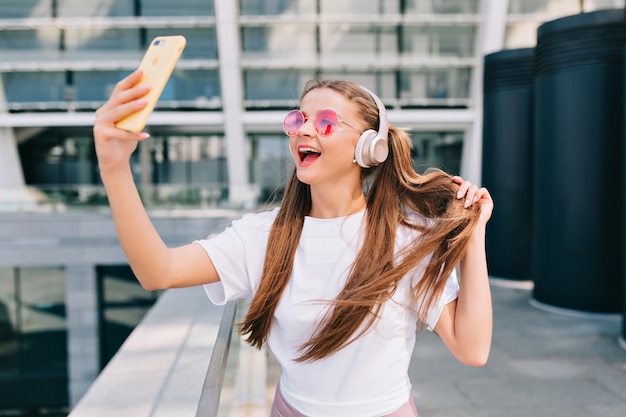 笑顔で踊る若い女性がスマートフォンで自分撮りをし、ヘッドフォンで音楽を聴く