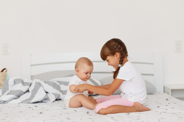 Smiling dark haired female kid with pigtails wearing casual clothing sitting on bed in light room, child holding baby's hands, spending time together.