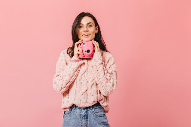 Smiling dark-haired lady in stylish sweater poses with pink camera on isolated background.