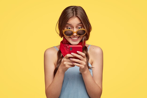 Smiling dark haired woman with cheerful expression, holds red mobile phone, happy to read text message, connected to wireless internet, isolated over yellow wall. People, technology, leisure