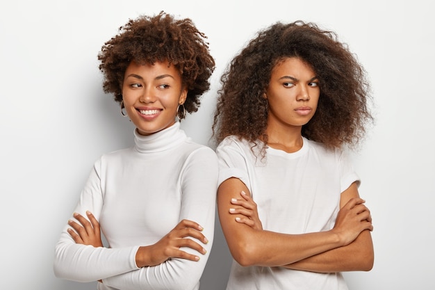 Smiling dark skinned woman keeps hands crossed, stand shoulder to shoulder with best friend who has gloomy upset expression, wear white clothes, express different emotions.