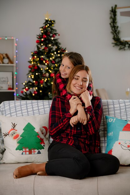 Smiling daughter hugs mother sitting on couch from behind and enjoying christmas time at home