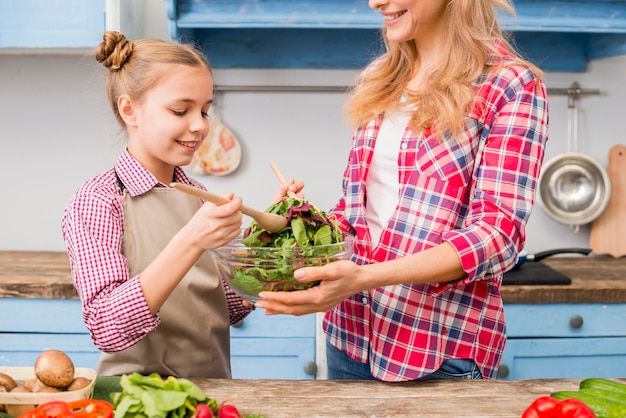 Улыбающиеся дочь и мать готовят листовой овощной салат на кухне