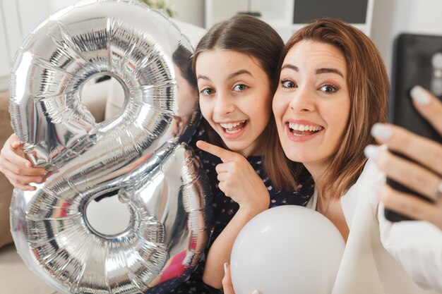 Улыбающаяся дочь и мать с воздушным шаром номер восемь в счастливый женский день, сидя на диване, делают селфи в гостиной