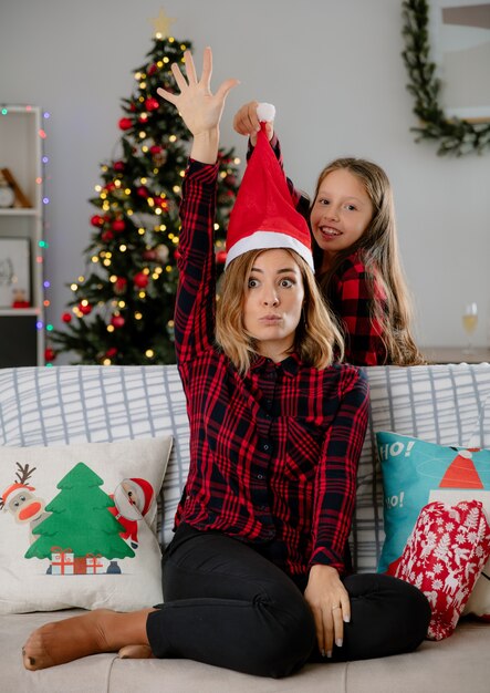 Smiling daughter puts santa  hat on mother  head raising hand up sitting on couch and enjoying christmas time at home