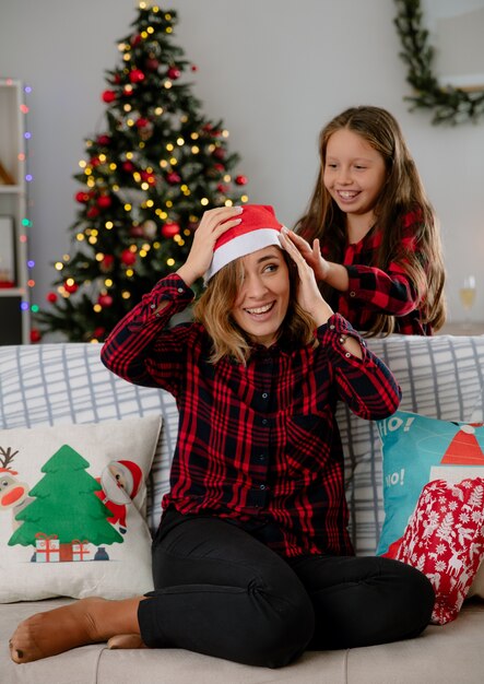 smiling daughter puts santa hat on mother head sitting on couch and enjoying christmas time at home