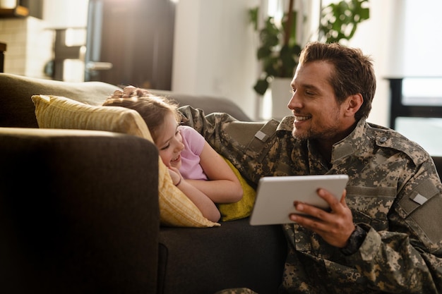 Улыбающаяся дочь отдыхает на диване и использует цифровой планшет со своим отцом-военным в гостиной