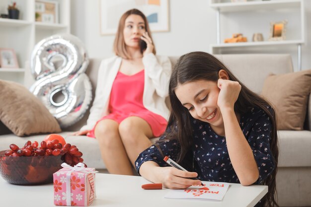 コーヒーテーブルの後ろの床に座っている笑顔の娘がプレゼントを持って幸せな女性の日にグリーティングカードに書き込みソファに座っている母親がリビングルームで電話で話します