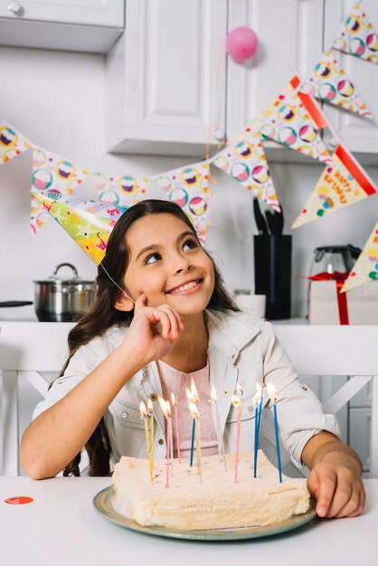 Smiling day dreaming girl sitting in front of birthday cake with illuminated candles