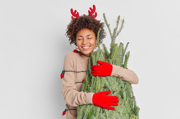 Восторженная улыбающаяся афроамериканка в оленьих рогах и перчатках с любовью обнимает зеленую елку, счастливая встречать Новый год дома, возвращается с рождественского уличного рынка, завернутая в гирлянду