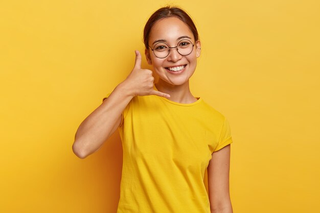 Smiling delighted Asian girl shows call me gesture, makes phone hand sign, has happy expression, healthy skin, wears spectacles and casual wear, isolated on yellow wall. Body language.