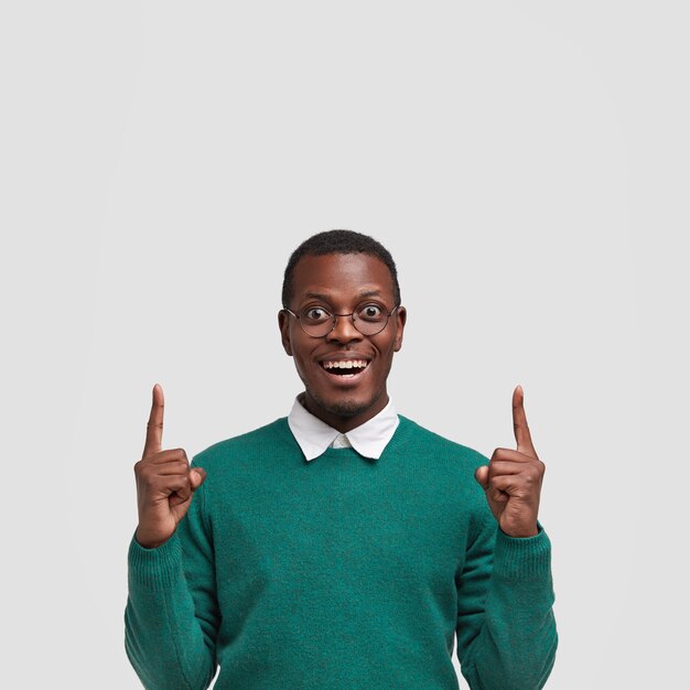 Smiling delighted black young man raises two fore fingers upwards, has glad facial expression, shows free space above head