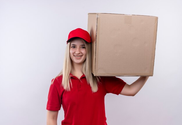 Smiling delivery young girl wearing red t-shirt and cap in dental brace holding big box on her shoulder on isolated white background