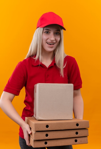 Smiling delivery young girl wearing red t-shirt and cap in dental brace holding box and pizza box on isolated orange background