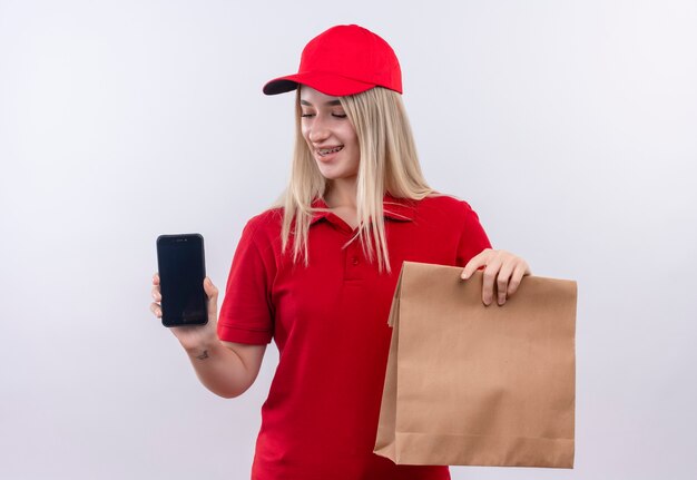 Smiling delivery young girl wearing red t-shirt and cap in dental brace holding phone and paper pocket on isolated white background