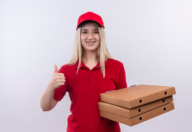 Smiling delivery young girl wearing red t-shirt and cap in dental brace holding pizza box her thumb up on isolated white background