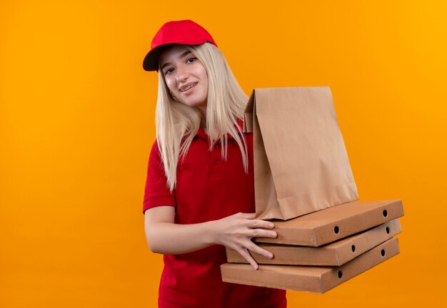 Smiling delivery young girl wearing red t-shirt and cap in dental brace holding pizza box and paper pocket on isolated orange background