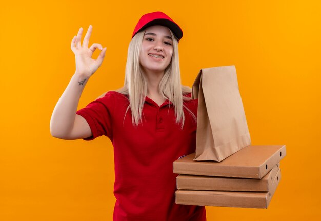Улыбающаяся молодая девушка из службы доставки в красной футболке и кепке в стоматологической скобе, держащая коробку для пиццы и бумажный карман, показывая жест на изолированном оранжевом фоне