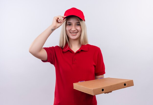 ピザボックスを保持している歯科ブレースで赤いTシャツとキャップを身に着けている笑顔の配達の若い女の子は、孤立した白い背景の上のキャップに彼女の手を置きます