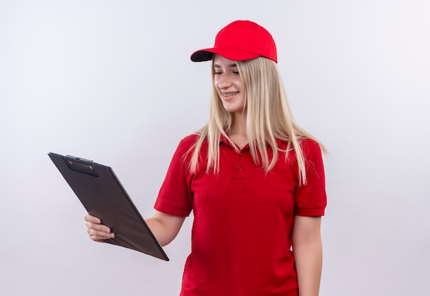 Улыбающаяся молодая девушка из службы доставки в красной футболке и кепке в стоматологической скобе смотрит в буфер обмена на изолированном белом фоне