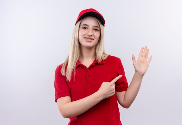 Smiling delivery young girl wearing red t-shirt and cap in dental brace points at hand on isolated white background