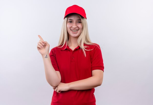 Smiling delivery young girl wearing red t-shirt and cap in dental brace points to side on isolated white background