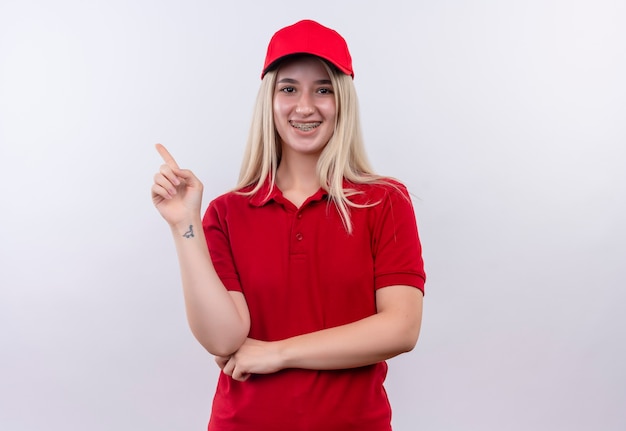 孤立した白い背景の上の側に歯のブレースで赤いTシャツとキャップを身に着けている笑顔の配達の若い女の子