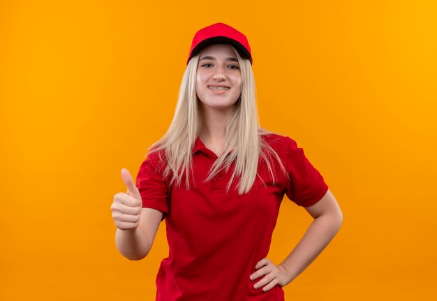 Улыбающаяся молодая девушка из службы доставки в красной футболке и кепке в стоматологической скобе положила руку на бедро, подняв большой палец на изолированном оранжевом фоне